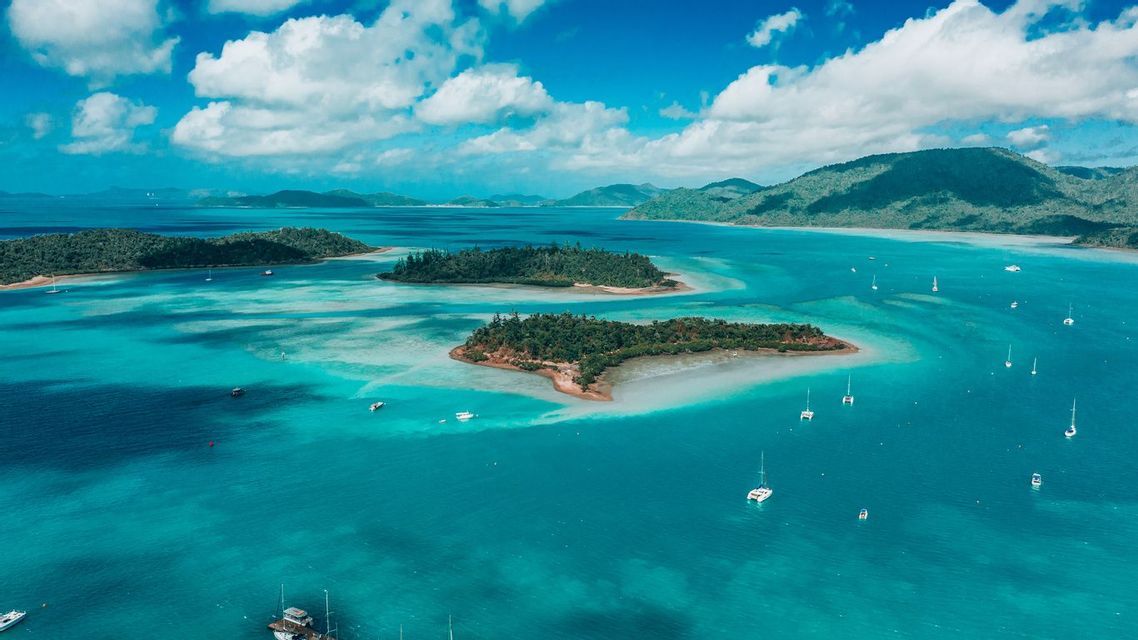 Veduta aerea di barche a vela e catamarani che galleggiano in acque turchesi, circondando piccole isole boscose sotto un cielo azzurro con nuvole.