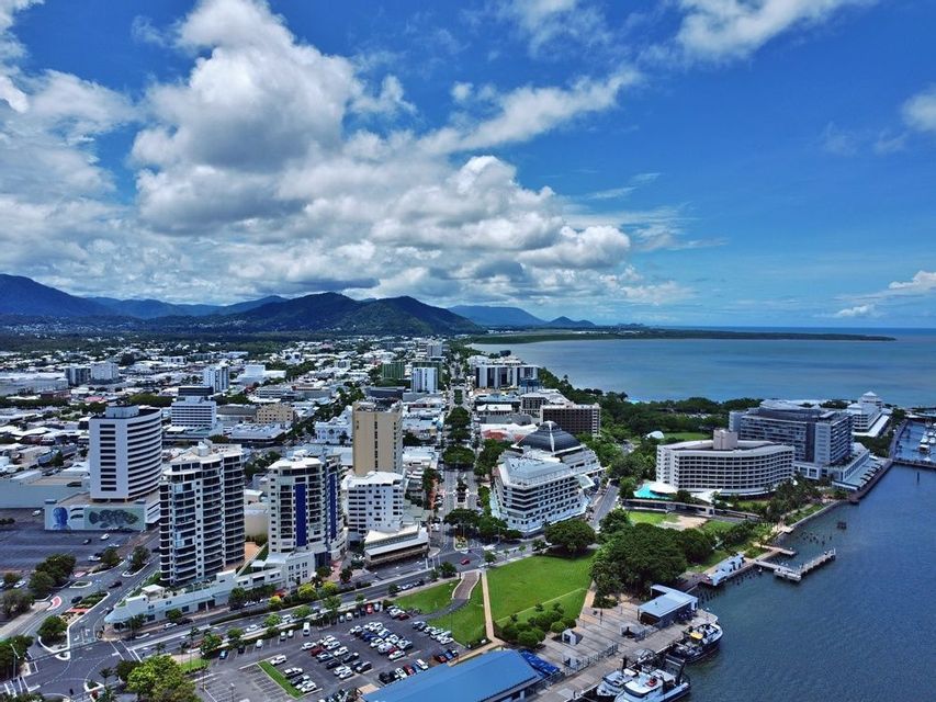 Vue aérienne d&#39;une ville côtière avec des bâtiments modernes, un port et des montagnes verdoyantes en arrière-plan sous un ciel bleu partiellement nuageux.