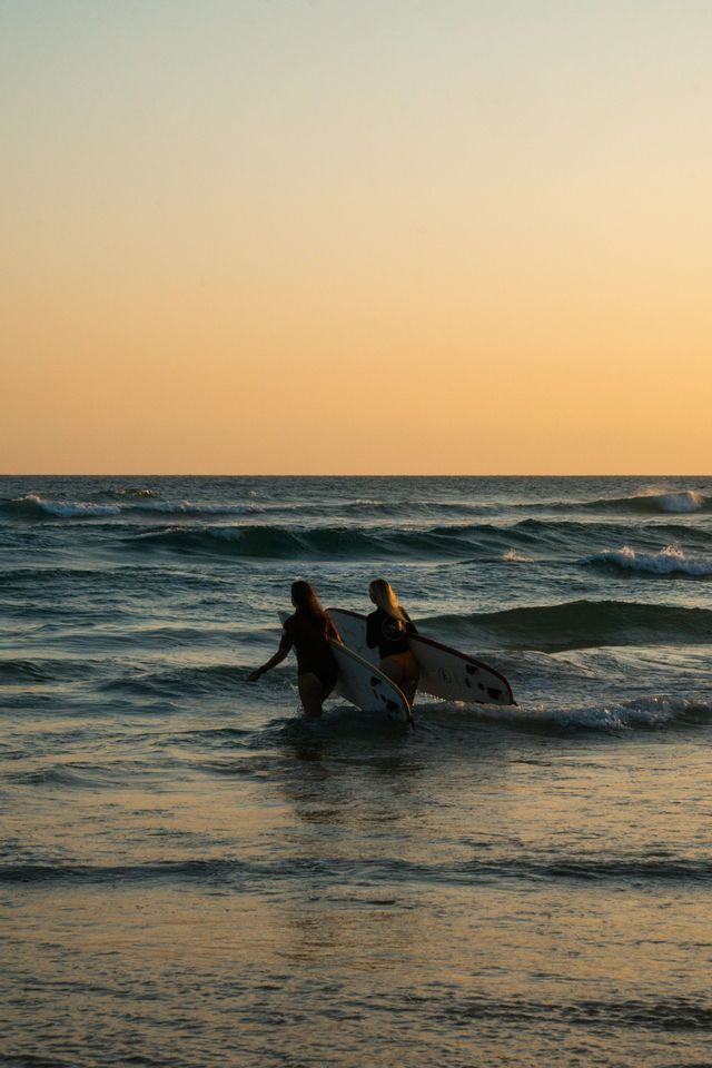 Due donne con tavole da surf camminano tra le onde dell'oceano sotto un caldo cielo al tramonto.