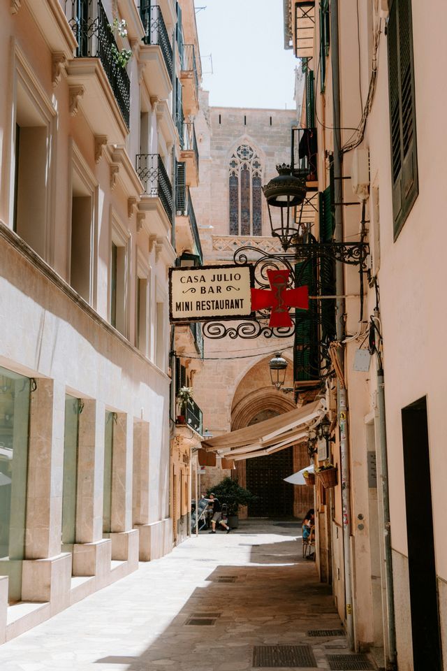 Un estrecho callejón soleado, con edificios históricos y un letrero de restaurante, lleva hacia una iglesia de piedra al fondo.