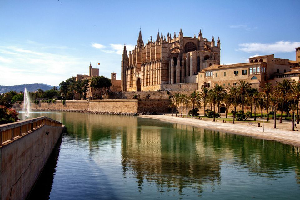 Una grande cattedrale gotica e gli edifici adiacenti si riflettono nell'acqua calma di un lago, con palme che costeggiano la riva.