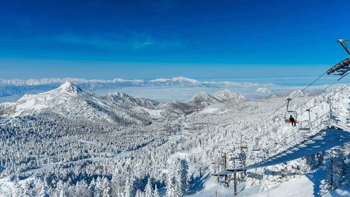 A chairlift ascends over a vast, snow-covered mountain landscape with pine forests and distant peaks under a clear sky.