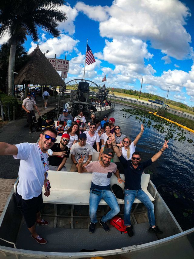 Un groupe WeRoad sourit et pose pour un selfie à bord d'un hydroglisseur sur l'eau sous un ciel bleu et nuageux.