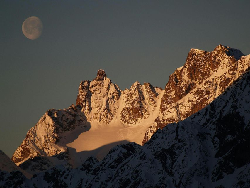 A large moon in a clear blue sky rises above snow-capped mountain peaks illuminated by the golden light of the sun.