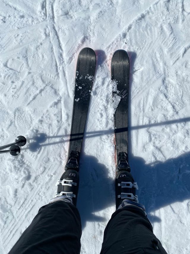 Vue à la première personne sur des skis et des chaussures de ski sur une pente enneigée et ensoleillée.