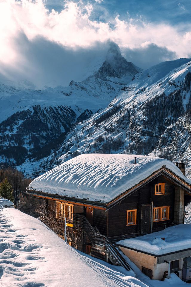 A wooden chalet with a snow-covered roof sits on the side of a mountain, with a spectacular peak visible behind it through the clouds.