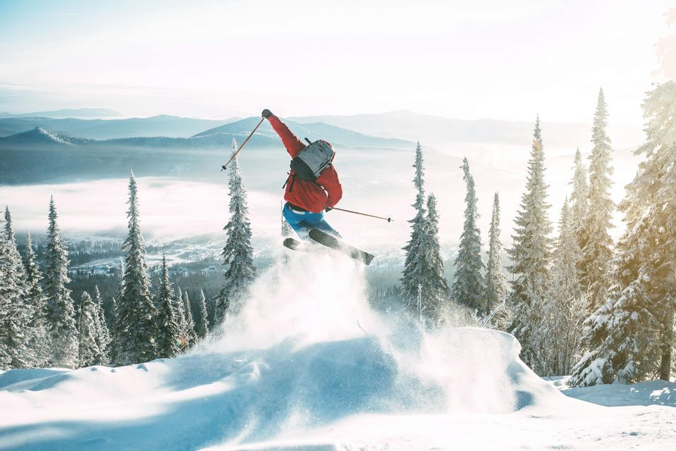 A skier in a red jacket jumps in mid-air, kicking up snow against a backdrop of snow-capped mountains and pine trees.