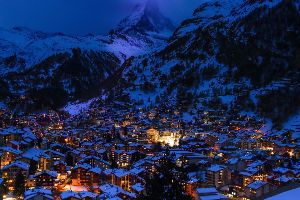 An illuminated village with snow-covered roofs sits in a mountain valley at sunset, with a towering, snow-capped peak in the background.