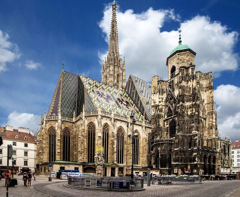 Una grande cattedrale gotica con un tetto di tegole colorate e decorate e un'alta guglia che si erge in una piazza cittadina sotto un cielo blu.