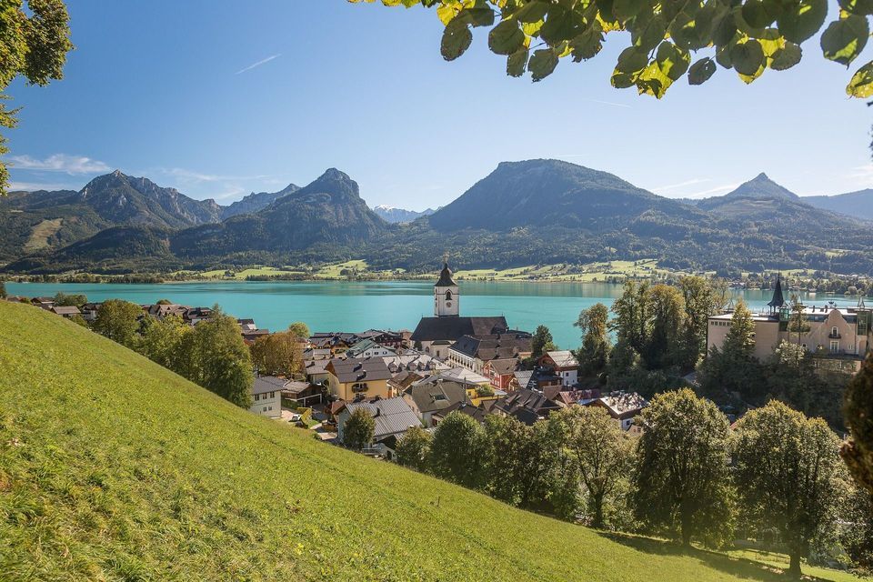 Un villaggio con un campanile si affaccia sulla riva di un lago turchese, con grandi montagne sullo sfondo, visto da una collina erbosa.