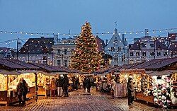 Un grande albero di Natale illuminato si erge al centro di una vivace piazza del mercato, piena di gente che curiosa tra le bancarelle al crepuscolo.