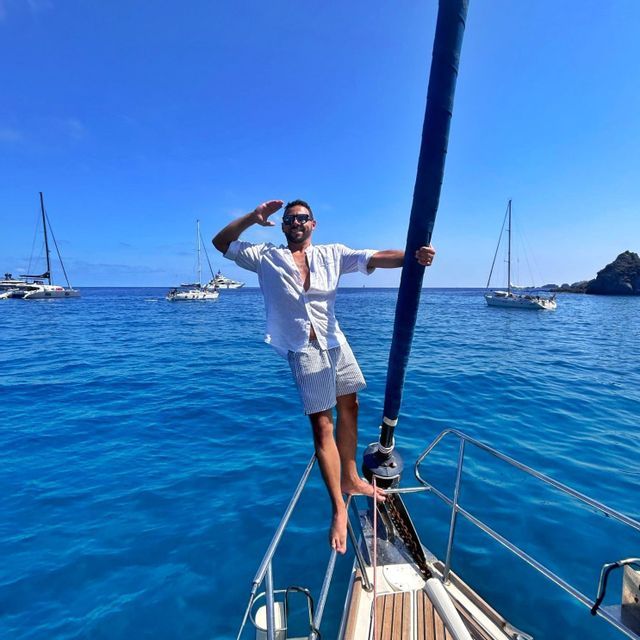 Ein lächelnder Mann mit Sonnenbrille steht am Bug eines Segelboots und hält den Mast, während es auf strahlend blauem Meer, umgeben von anderen Booten, segelt.