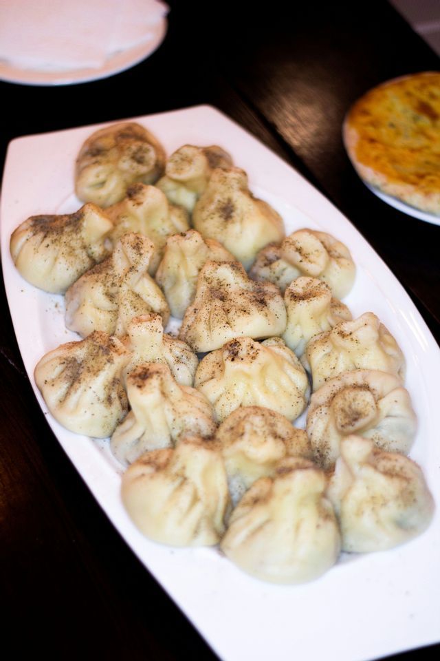 A white plate of steamed ravioli, sprinkled with black pepper, sits on a dark wooden table.