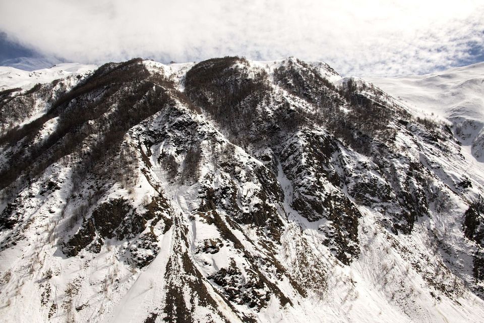 A steep, rugged mountain, covered in snow, with patches of rock and bare trees visible under a partly cloudy sky.