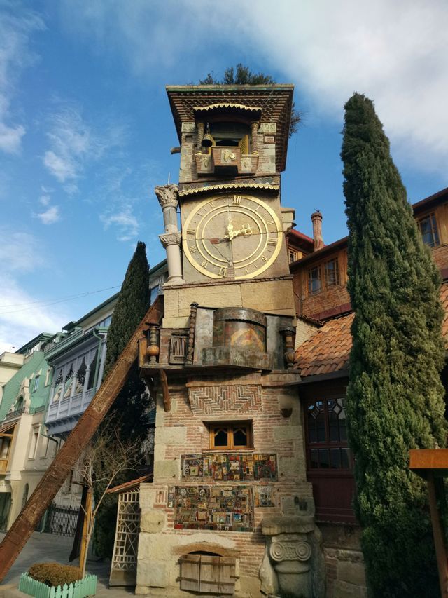 Una caprichosa torre del reloj inclinada con una gran esfera dorada se alza en una calle de la ciudad junto a un alto ciprés.