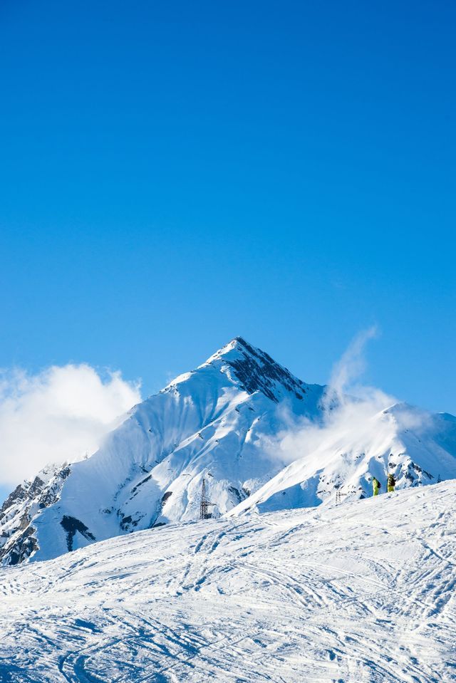 A vast, snow-covered mountain peak stands against a clear blue sky, with a WeRoad group trip skiing on the slopes below.