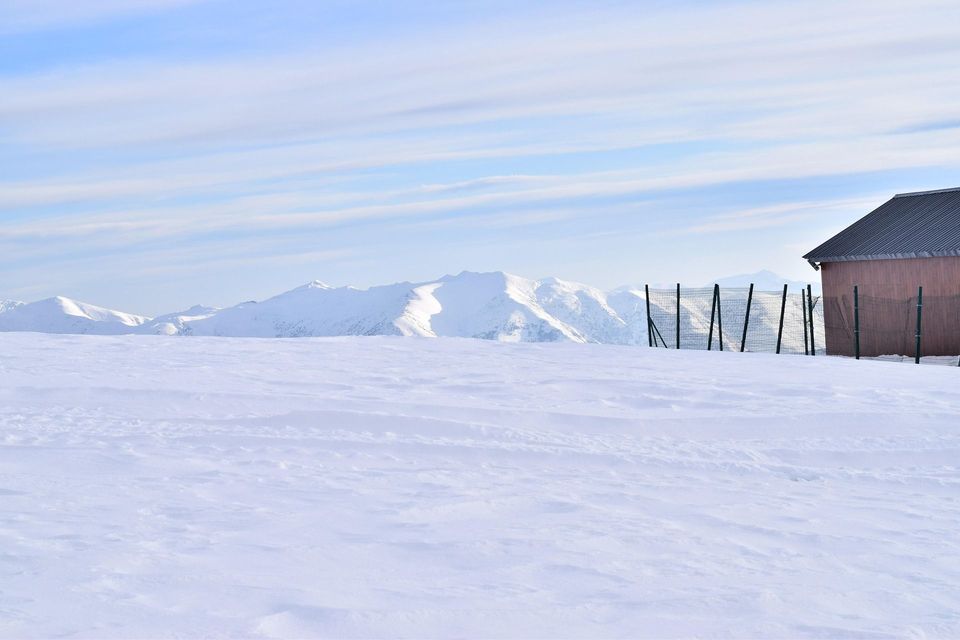 A wide snowy landscape with a wooden cabin and a fence on the right, and snow-capped mountains in the background.