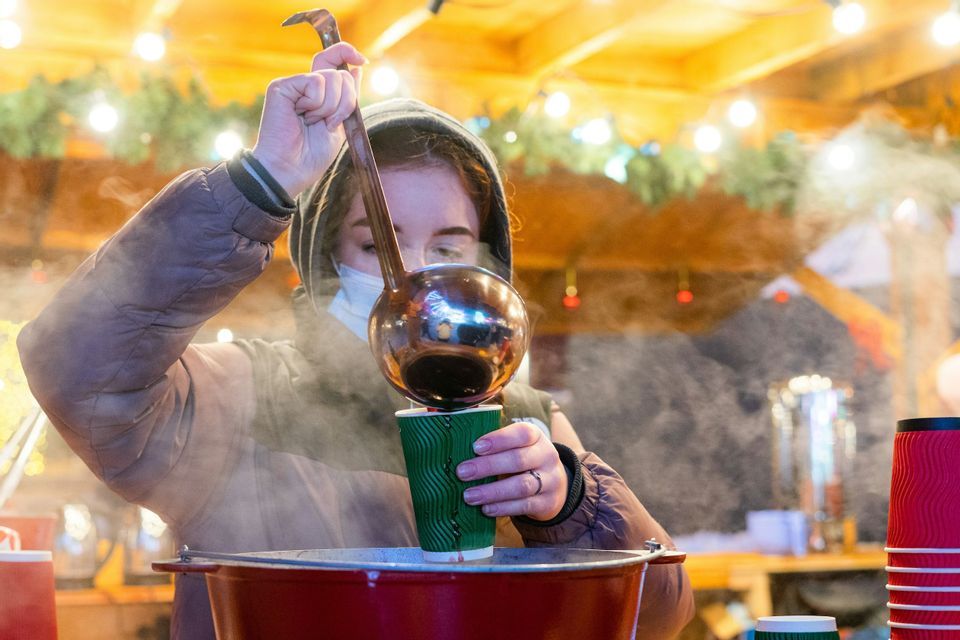 A person wearing a hooded jacket and face mask uses a large ladle to pour a steaming drink into a green cup from a large pot.