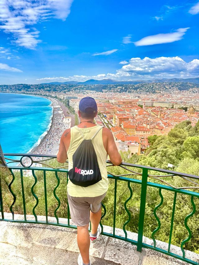 Un hombre con una mochila WeRoad observa desde un mirador una ciudad costera con una playa y mar turquesa.