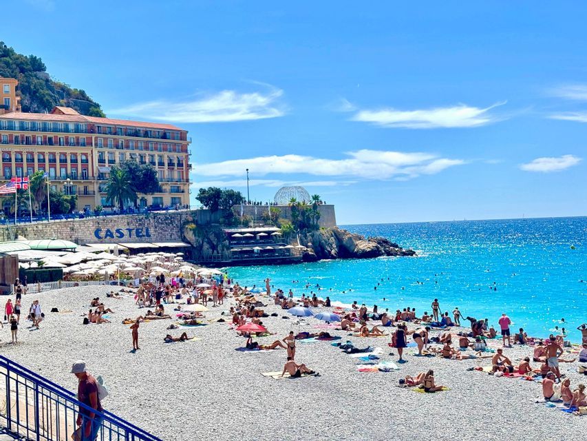 Vista de una concurrida playa de guijarros donde la gente toma el sol y nada en aguas turquesas bajo un cielo azul despejado.