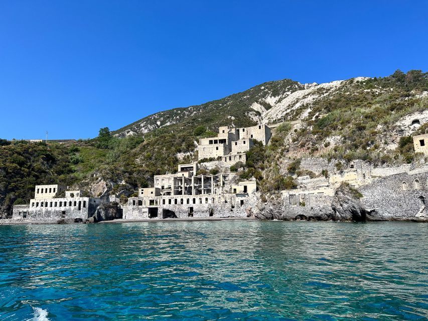 Edifici in pietra abbandonati incastonati in un verde pendio, sul bordo di un mare turchese sotto un cielo azzurro e limpido.