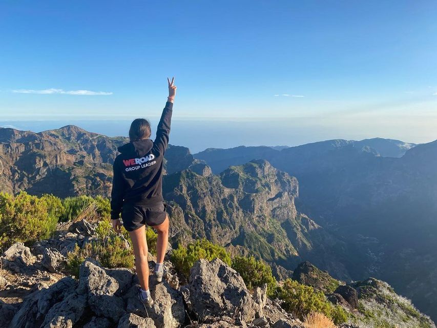 A WeRoad group leader stands on a rocky mountain summit, making a peace sign while overlooking a vast mountain range.