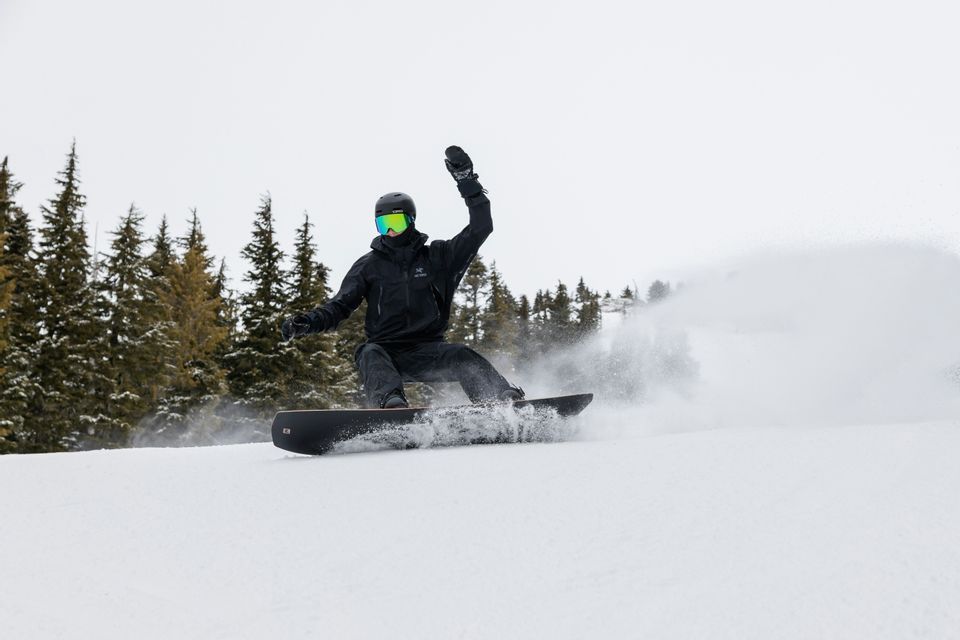 Uno snowboarder con attrezzatura nera scende tracciando curve su un pendio innevato, sollevando neve con una mano alzata.