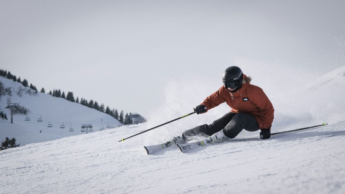 Una persona con una giacca arancione e un casco nero scia su un pendio innevato, sollevando neve fresca, con uno skilift sullo sfondo.