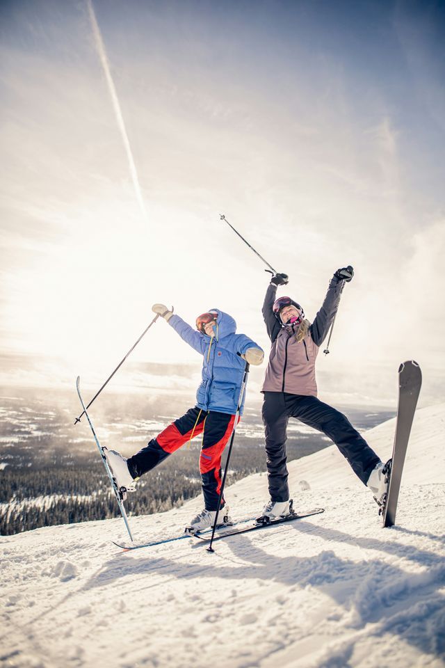 Due persone di un viaggio di gruppo WeRoad posano con le braccia alzate su un pendio innevato, con sci e snowboard, sotto un sole splendente.