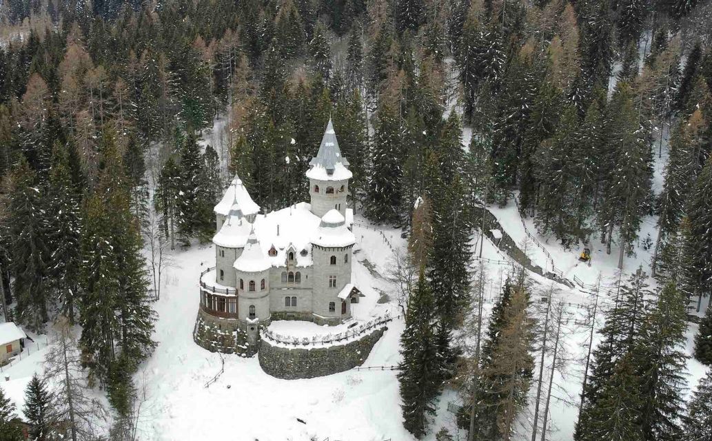 Una vista aerea di un castello di pietra con tetti e torrette innevati immerso in una fitta foresta di pini.
