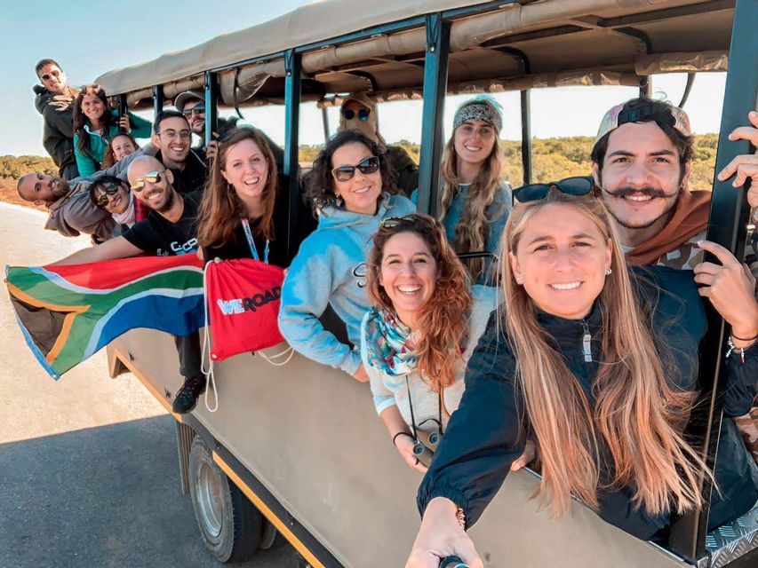 Eine lächelnde WeRoad-Gruppe posiert in einem offenen Safari-Fahrzeug für ein Selfie und zeigt eine südafrikanische Flagge.