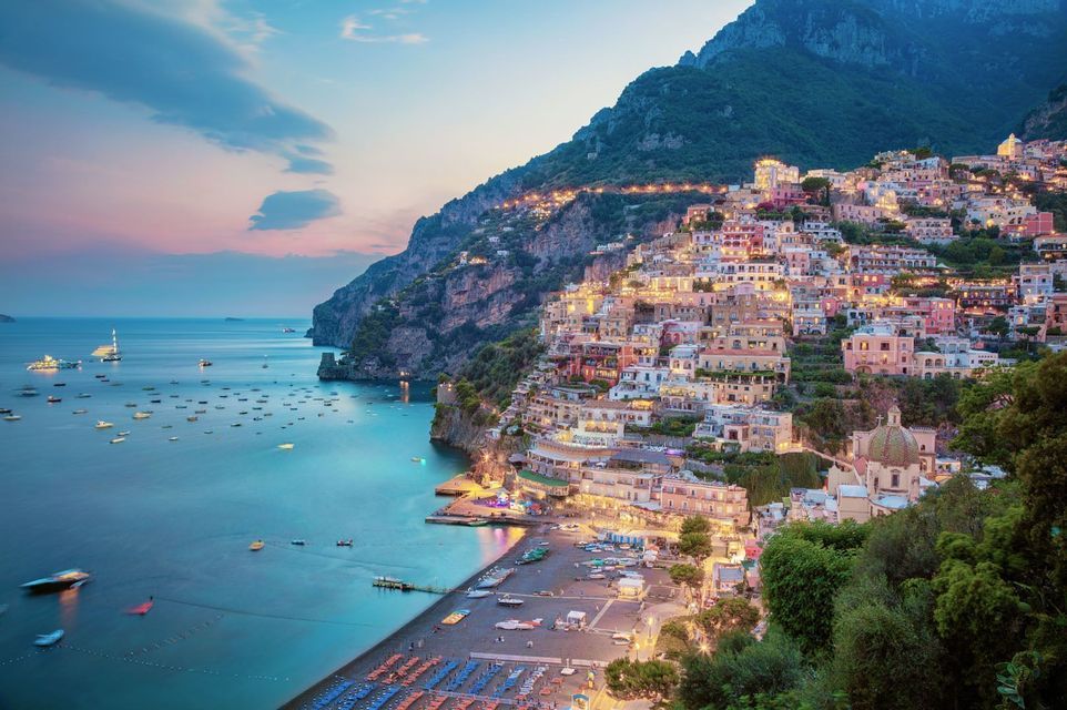 A colorful, illuminated town built on a steep cliffside, overlooking a calm sea with many boats at dusk.