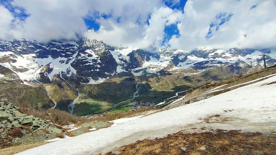 Une vue panoramique d'une chaîne de montagnes enneigées surplombant une vallée verdoyante avec un petit village, observée depuis une pente enneigée sous un ciel nuageux.