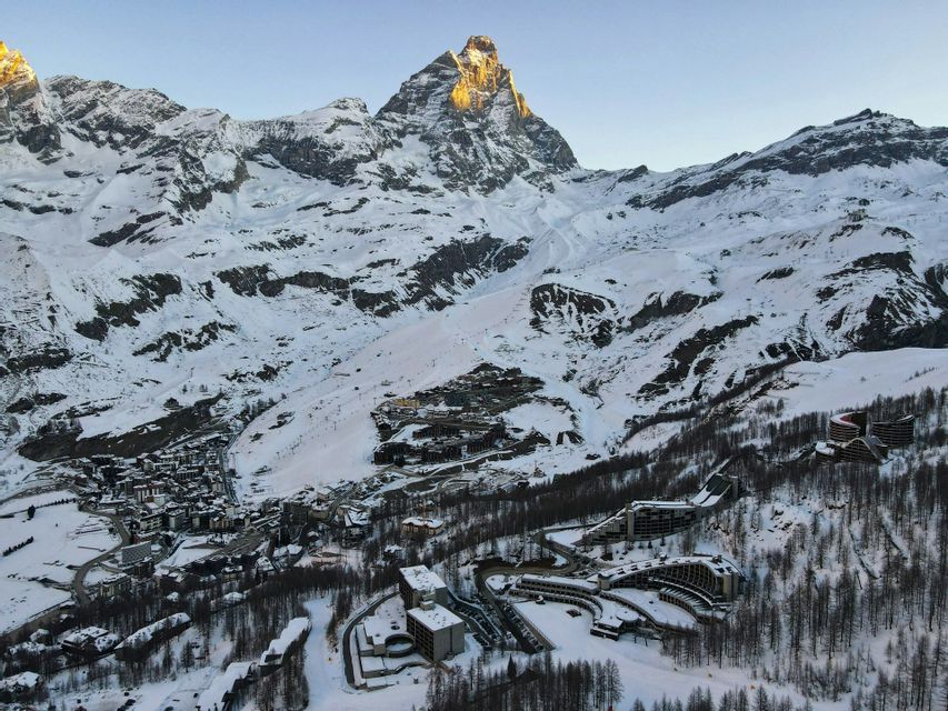An aerial view of a ski resort village nestled in a snowy valley at the foot of a large mountain peak lit by the sun.