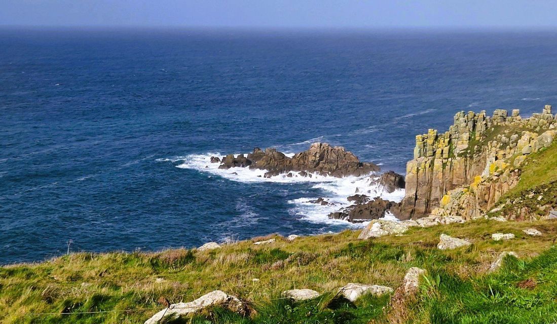 Una vista da una scogliera erbosa che si affaccia su un oceano blu profondo con onde che si infrangono contro formazioni rocciose costiere.