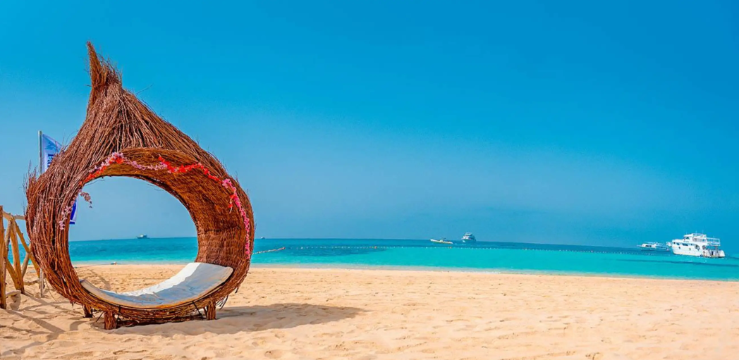 Un transat en osier façon nid, avec un coussin blanc, est installé sur une plage de sable fin, face à des eaux turquoise sous un ciel bleu clair.