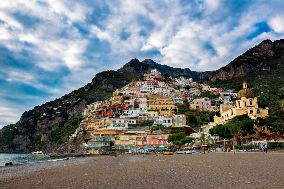 Des maisons colorées en cascade le long d'une pente raide menant à une plage de sable et à la mer, sous un ciel nuageux.