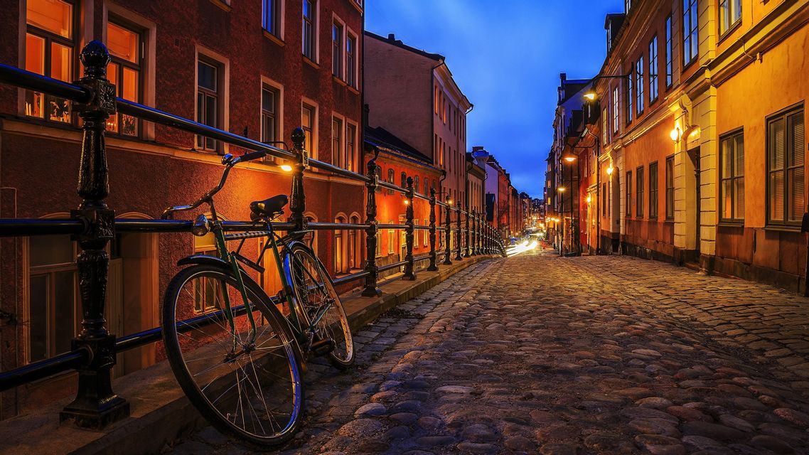 Una bicicletta è appoggiata a una ringhiera su una stretta strada acciottolata fiancheggiata da edifici illuminati al tramonto.
