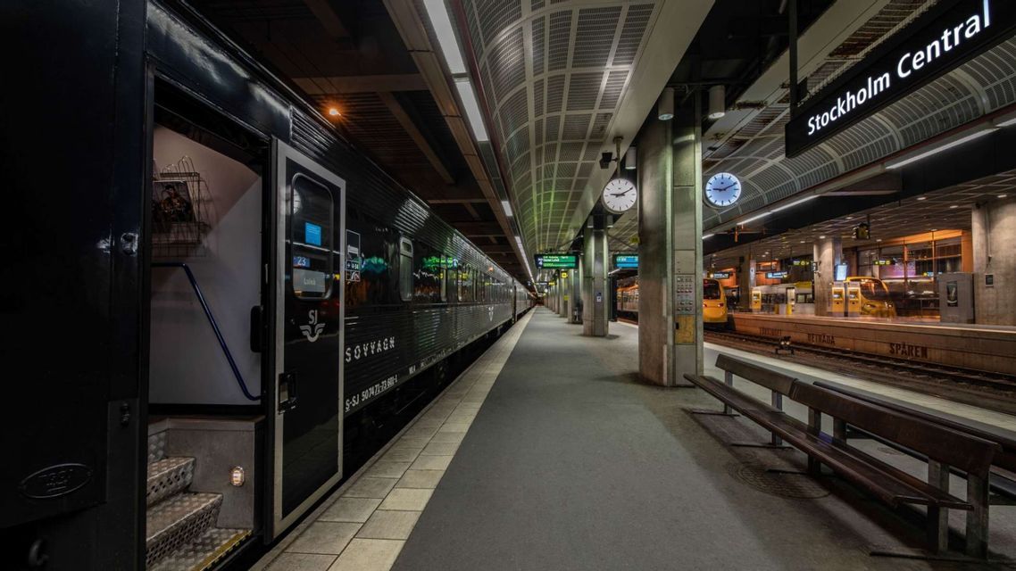Un treno letto nero con una porta aperta si trova su una banchina vuota e illuminata in una moderna stazione ferroviaria di notte.
