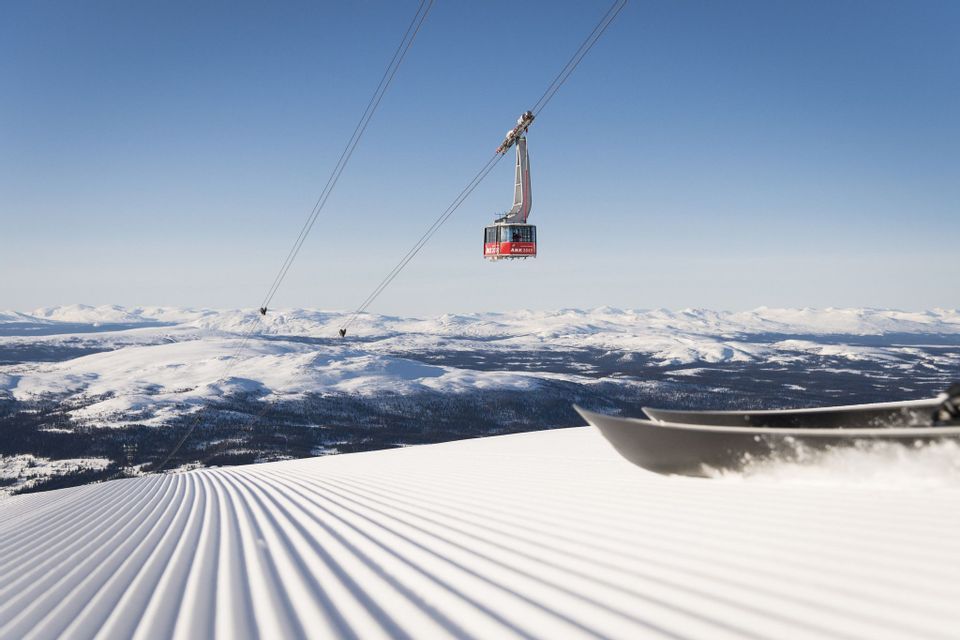 Una funivia viaggia sopra una pista da sci appena battuta, con le punte di uno snowboard in primo piano e montagne innevate sullo sfondo.