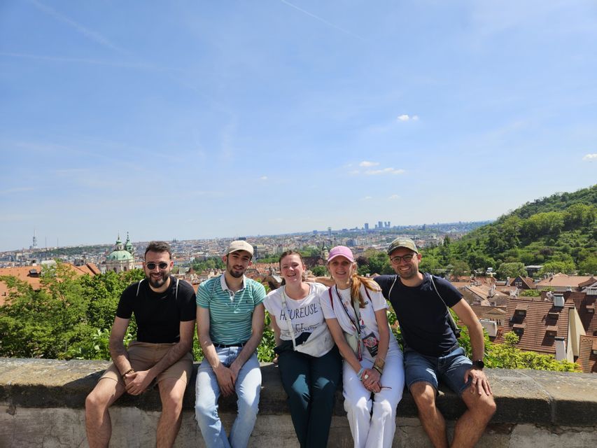 Fünf Freunde einer WeRoad-Gruppe sitzen lächelnd auf einer Steinmauer und genießen den Blick auf eine europäische Stadtlandschaft unter klarem blauem Himmel.