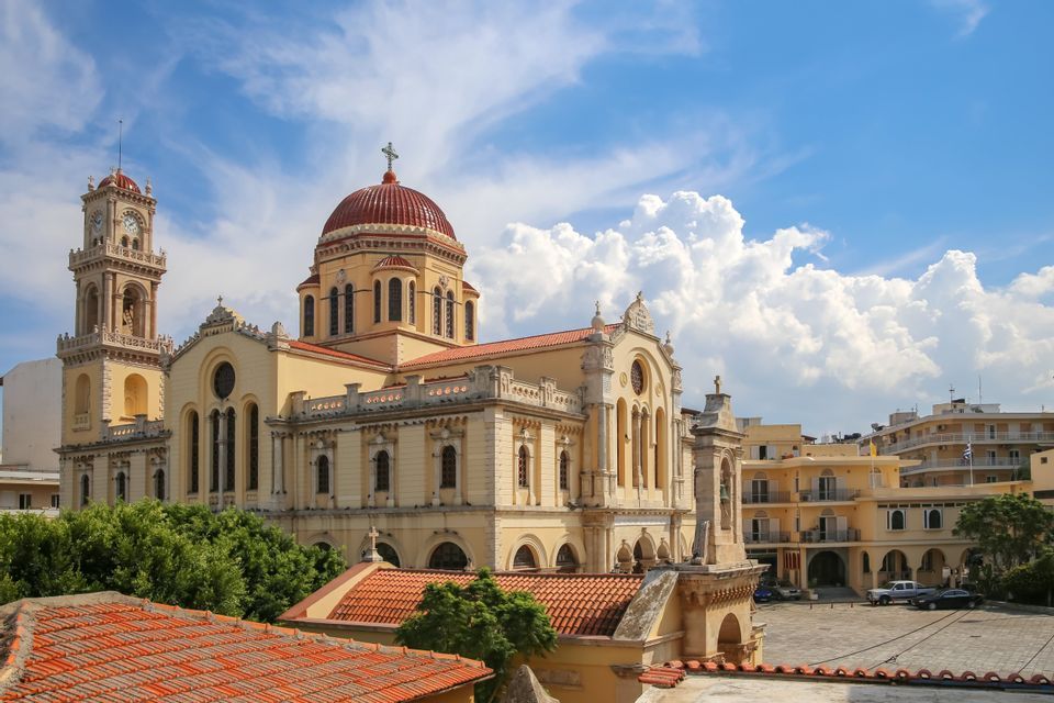 Una grande cattedrale gialla con una cupola rossa e una torre dell'orologio si erge in una piazza cittadina sotto un cielo azzurro nuvoloso.