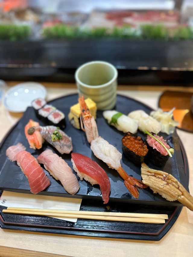 Una varietà di sushi fresco, inclusi nigiri e maki, disposti su un piatto di portata nero con una tazza da tè e bacchette.