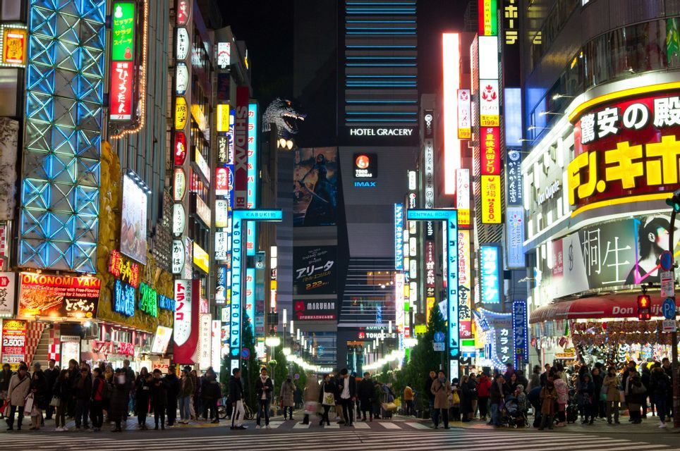 Una folla di persone si trova su un attraversamento pedonale cittadino di notte, circondata da alti edifici coperti da insegne al neon colorate e una grande testa di Godzilla.