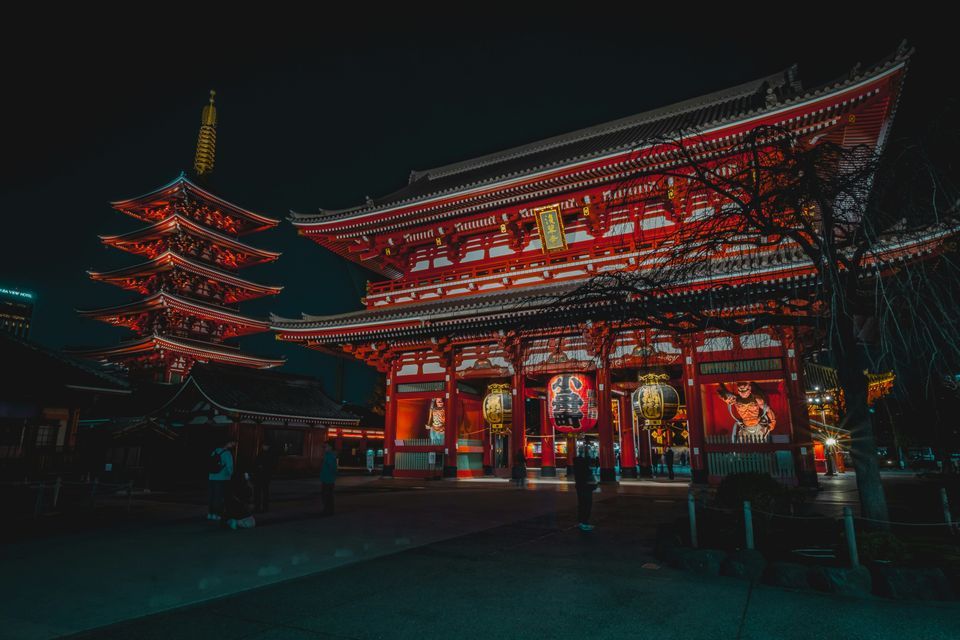 Un grande tempio giapponese rosso e una pagoda a cinque piani illuminati di notte, con persone nel cortile.