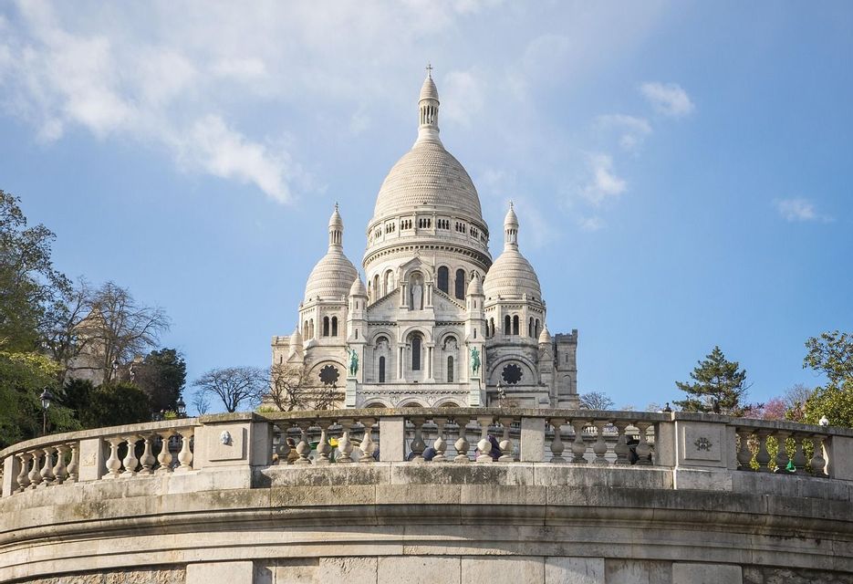 Une grande basilique en pierre blanche avec plusieurs dômes, vue de derrière une balustrade en pierre sous un ciel bleu partiellement nuageux.