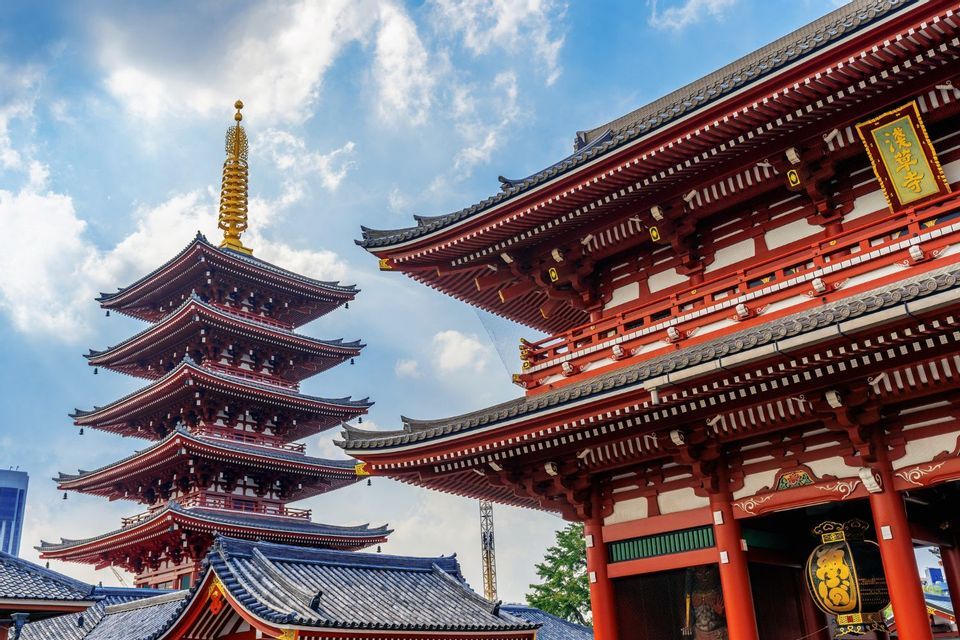 Una pagoda giapponese rossa a più piani e un tradizionale portale del tempio si ergono contro un cielo blu parzialmente nuvoloso.