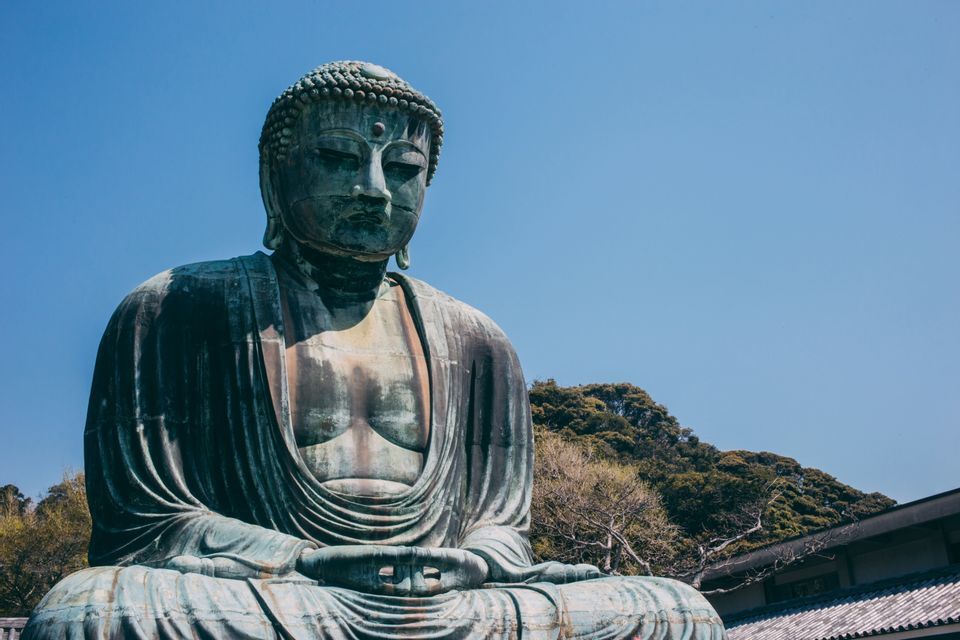 Una grande statua in bronzo di un Buddha seduto all'aperto contro un cielo azzurro limpido, con una collina verde sullo sfondo.