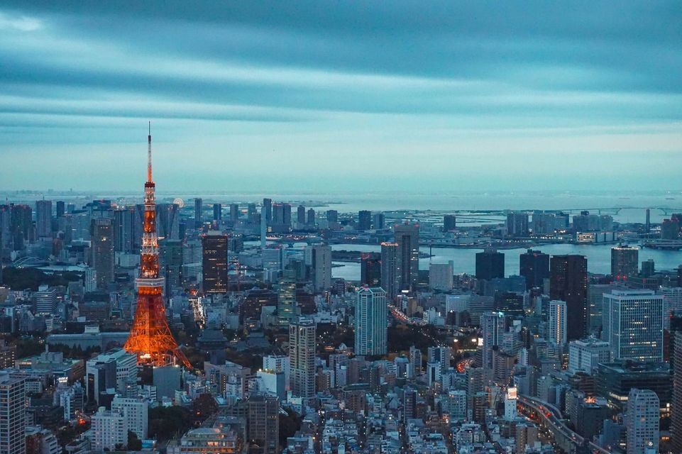 La Torre di Tokyo illuminata di arancione si staglia contro il denso paesaggio urbano al tramonto, con una baia visibile sullo sfondo.