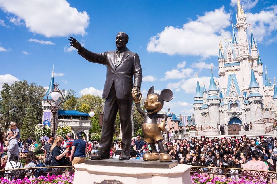 Une statue en bronze d'un homme tenant la main d'un personnage souris devant un grand château de conte de fées entouré d'une foule.